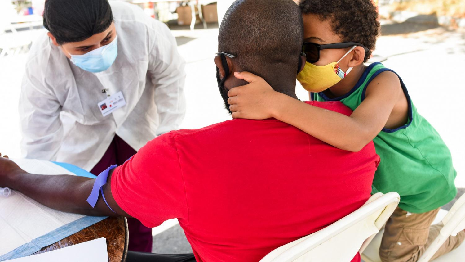 A volunteer healthcare worker at the Fruitvale COVID Testing Event draws blood from an African American man. His young son hugs him and provide reassurance