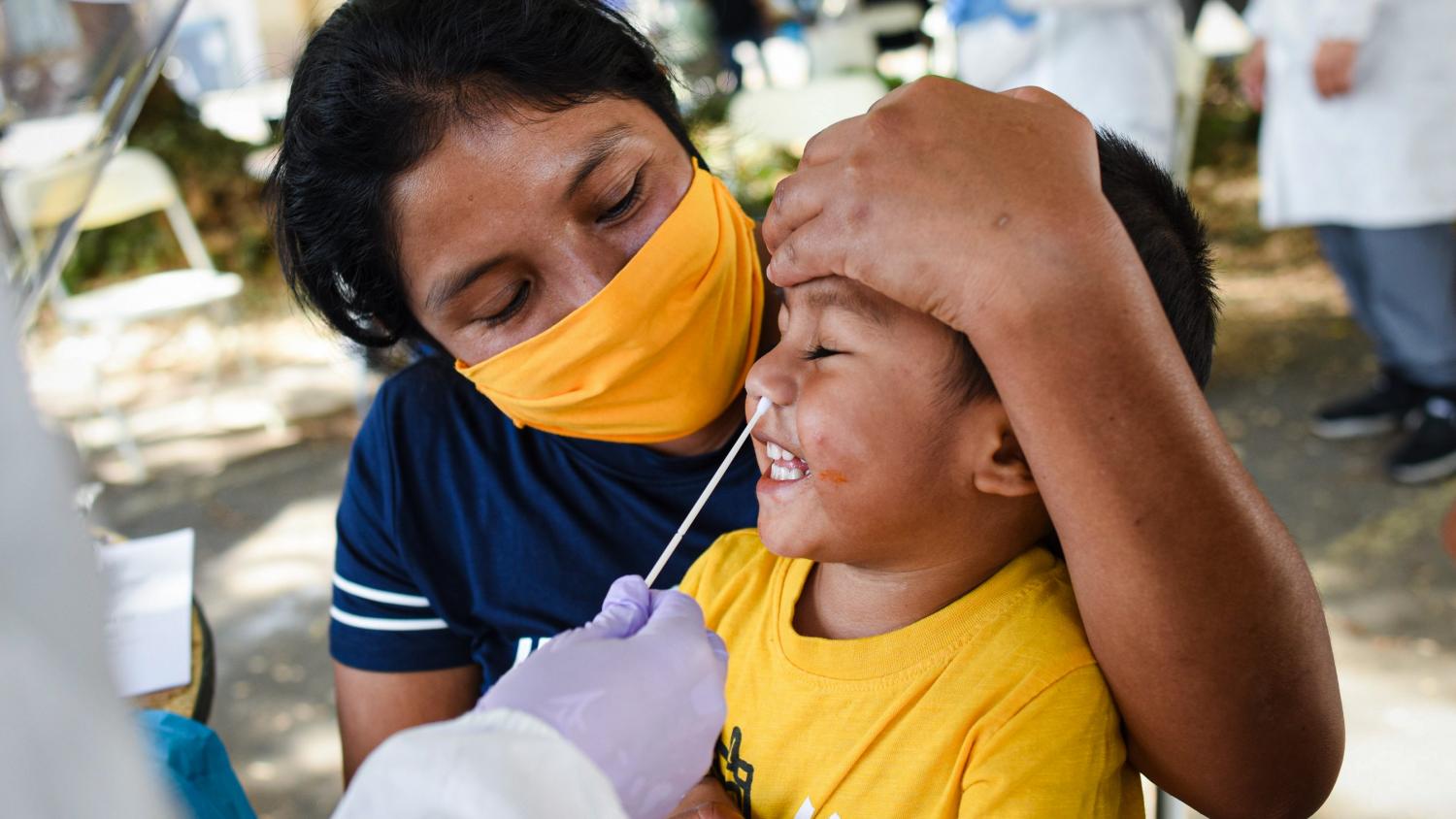 A volunteer healthcare worker swabs the nose of a young Latino boy. He is smiling while his mom hugs hugs him and holds his head.
