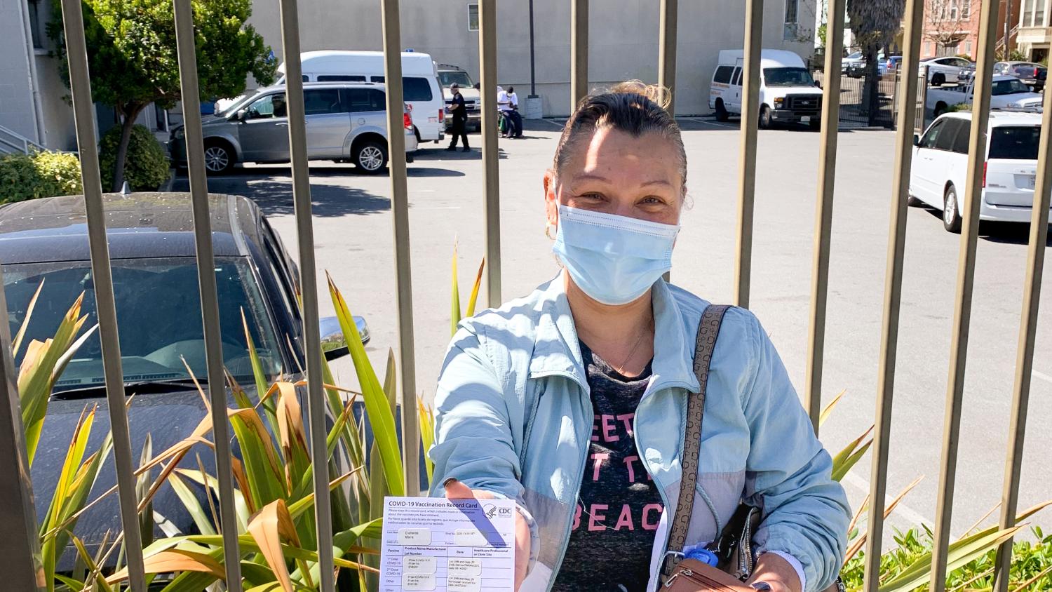 A Latina woman wearing a mask, stands in front of a gate  while showing her proof of vaccination card.