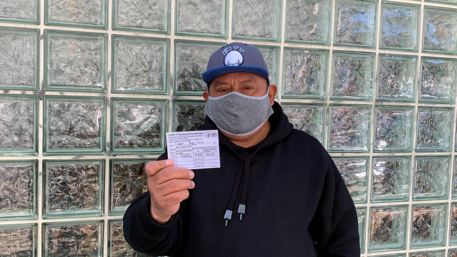A Latino man wearing a mask, stands in front of a building, holding his proof of vaccination card